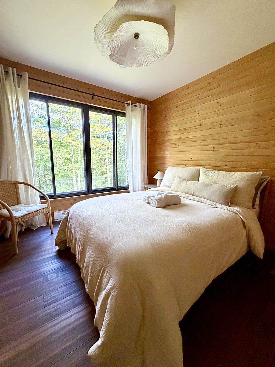 La chambre claire : lit en lin, rideaux blancs et fenêtre sur les arbres.