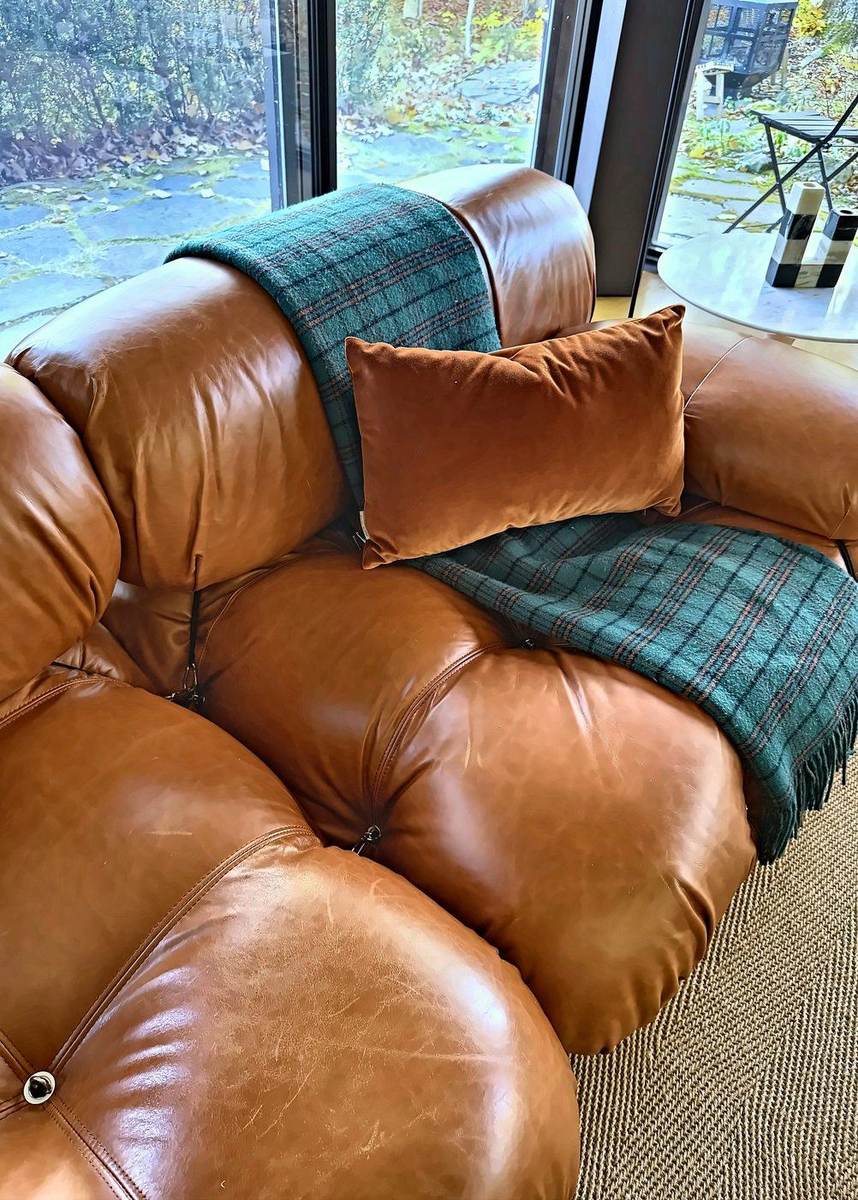 Le canapé en cuir cognac patiné, un plaid écossais vert et un coussin rouille.